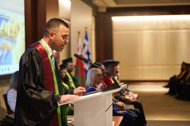 George Ntemos addressing the ceremony from the lectern, with flags behind him.
