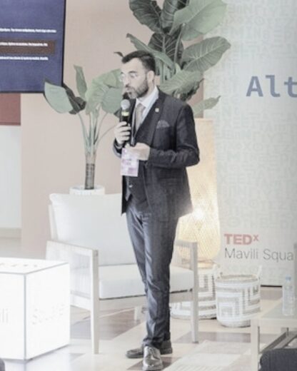 George Ntemos standing on the TEDxMaviliSquare stage, speaking into a microphone.