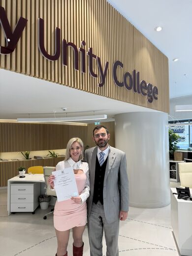 George Ntemos with a graduate holding her degree certificate, beneath the City Unity College sign.